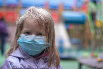 Portrait of a cute blonde little girl wearing a Medical Disposable Face Mask, playing on a playground during quarantine. Stay at home. Isolation. Coronavirus and Air pollution concept.