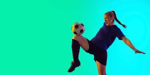 Young female soccer or football player with long hair in sportwear kicking ball for the goal, training on gradient background, neon. Concept of healthy lifestyle, professional sport, motion, movement.