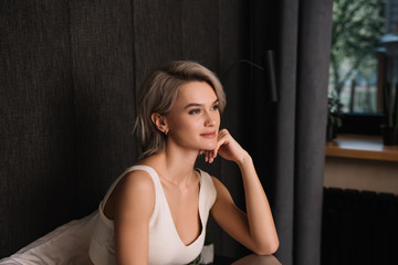 dreamy, positive woman touching face and looking away while sitting in bedroom