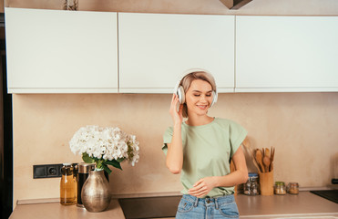 happy young woman listening music in wireless headphones in kitchen with closed eyes