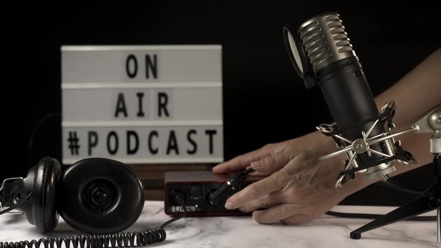 Podcast studio set with microphone, headphones, console and &ldquo;on air&rdquo; sign. A person connects cables, turns on console and sign. All over marble table and black background.
