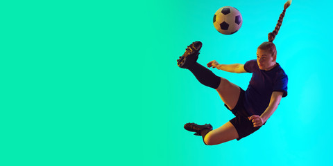Young female soccer or football player with long hair in sportwear kicking ball for the goal in jump on gradient background, neon. Concept of healthy lifestyle, professional sport, motion, movement.