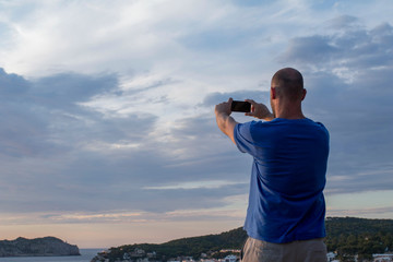 Fototapeta na wymiar Wanderlust concept of a man with his mobile phone at sunset in Sant Elm beach bay in Mallorca. The man is taking pictures at sunset
