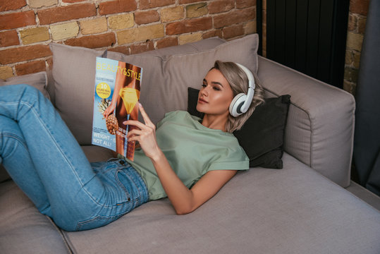 high angle view of young woman in wireless headphones reading beauty and style journal while relaxing on sofa at home