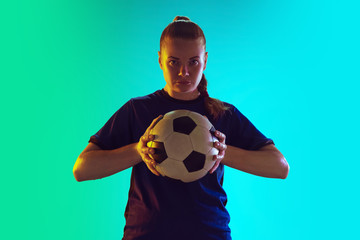 Young female soccer or football player with long hair in sportwear holding ball, posing confident on gradient background, neon. Concept of healthy lifestyle, professional sport, motion, movement.