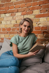 attractive young woman smiling at camera while sitting on sofa with open book