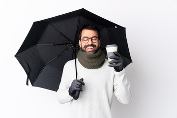 Caucasian handsome man with beard holding an umbrella and a coffee to take away over isolated white wall