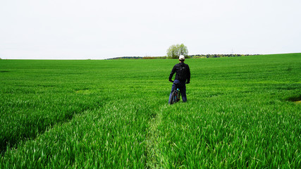 A man in a black sports jacket and a white cap on a bicycle among the green grass looks forward
