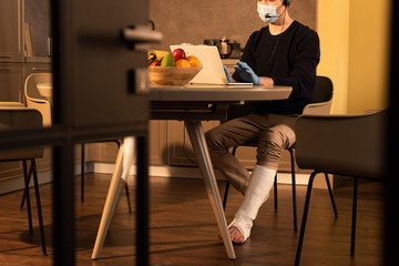 Selective focus of man in medical mask and plaster bandage on leg using headset and laptop in kitchen