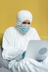 Fototapeta na wymiar Selective focus of man in hazmat suit and medical mask using laptop on bed