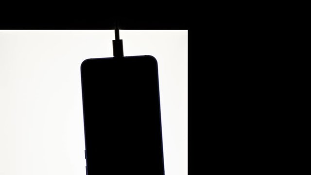silhouette of black smartphone hanging with usb charge cable on white and black blank area. communication and nomophobia concept, phone addiction