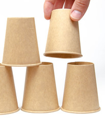 Brown paper cup for coffee, tea, a drink from environmental materials on a white background. The male hand builds a pyramid of cups in macro and overall plan. Place for text on the left and right.
