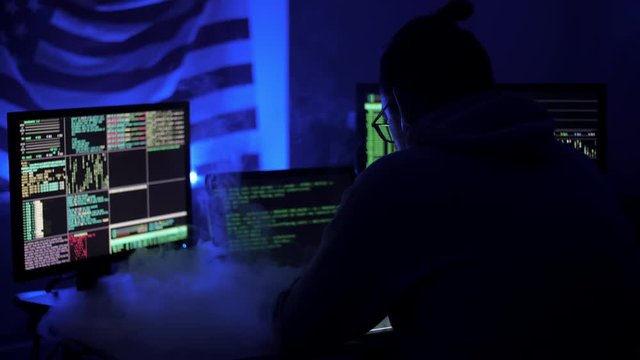 Programmer hard work in home to late night on computer. Hacker code blinks of the monitor screen. Man exhales smoke from a cigarette or vape and writes code and programs
