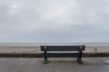 Image of a lonely black bench in England facing the sea with a long sand patch and stretch go pebbles going towards the water ready for someone depressed to sit on the lonely empty chair