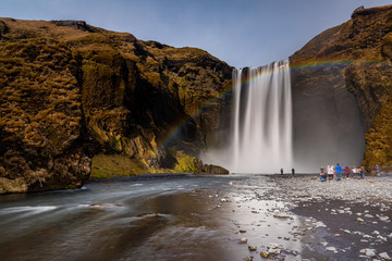 Skogafoss waterfall in South Iceland with a beautiful rainbow. Tourists admiring the waterfall. Popular and famous tourist attraction.