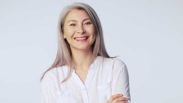 A friendly positive old mature woman with long gray hair wearing formal business clothes is laughing while looking to the camera standing isolated over white background in studio