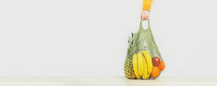 Delivery of grocery. Hand holds mesh shoper bag with fresh fruit and vegetables green produce. Eco friendly lifestyle and shopping. Healthy eating vegetarian, zero waste concept. Copy space. Banner