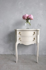 Room interior with bedside table on the gray loft wall. Glass vase with pink peonies on a Baroque table. Vintage furniture.