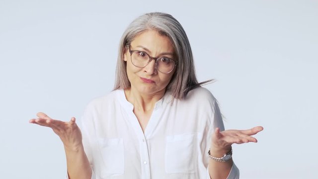 A confused disappointed old mature woman with long gray hair wearing glasses is raising her hands isolated over white background in studio