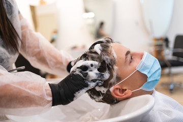 Profile of a young man with a protective mask leaning on a pike while washing his head in a salon