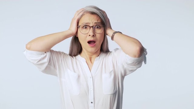 A shocked old mature woman with long gray hair wearing glasses is clutching her head isolated over white background in studio