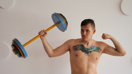 Young man with naked body and tattoo on chest lifting multicolored barbell against white patterned...