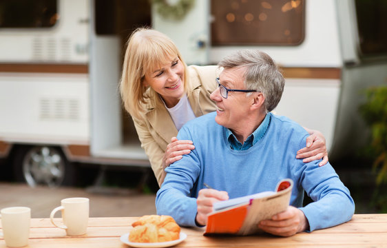 Mature Woman Hugging Her Husband While He Solving Crossword During Breakfast At Campsite