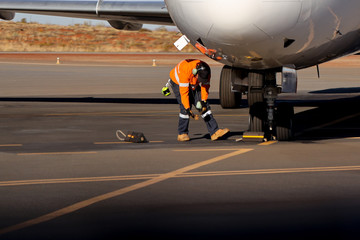 Safety practices aircraft worker wearing long sleeve hi-vis tape safety visible shirt wearing earmuffs glove kneeling double safety checking black rubber wheels chock placing under airplane wheels 