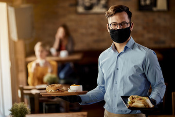 Happy waiter serving food while wearing protective face mask in a pub.