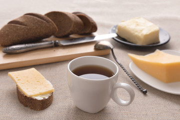 Sliced bread and knife on a cutting board. Butter, cheese, a cup of coffee on a sackcloth. Side view breakfast brunch on table concept