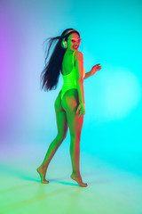 Beautiful young girl dancing with wireless headphones on gradient studio background in neon light. Woman in fashionable bodysuit. Facial expression, summer, weekend, beauty, resort concept. Vacations.