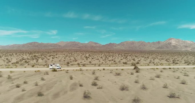 AERIAL - A brown camper van in the distance drives from left to right through the image while being passed by a small black car in the middle of the Mojave desert on a clear and blue day