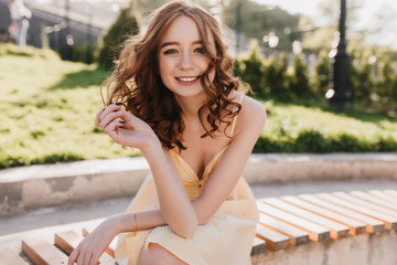 Naklejka premium Graceful ginger girl in yellow dress sitting in park. Lovely red-haired curly woman posing with inspired smile on nature background.