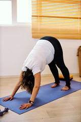 Middle aged pregnant woman standing on the mat at home in the morning in Downward-Facing Dog yoga posture.