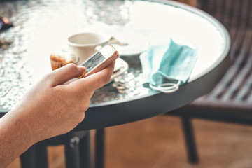 New normality. Drinking coffee on a terrace with the bill and the mask on the table, while looking at the phone.