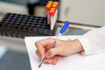 Doctor's hand writing test results from patient's blood test tubes on the white paper by pen in the laboratory.