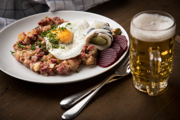 Norddeutscher Hamburger Labskaus mit Rindfleisch Corned Beef, Rote Beete, Kartoffeln, Saure Gurken, Spiegelei, Rollmops und Bier auf Holztisch