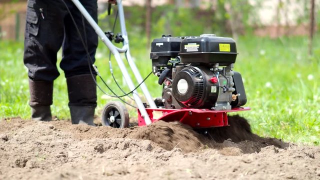 Man working with garden tiller engine in 4k VIDEO. Cultivator machine ploughing and shredding soil with blades on field at spring. Modern farming, technology agriculture. Close-up.