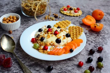 Healthy and tasty breakfast. Yogurt with granola and berries, waffles, nuts and peaches on a gray background.