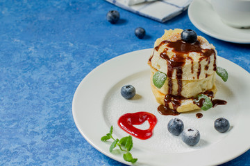 Japanese soft pancakes with blueberries, hot chocolate and sprinkled with icing sugar on a round white plate. Blue background. Copy space.