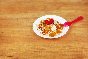 Delicious tiny pancakes with strawberries and butter lie on a white plate on a wooden background with space for text, next to a red spoon. Breakfast for children, trendy Breakfast, trendy food