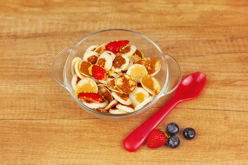 Tiny pancakes with blueberries and strawberries and yellow sprinkles in a glass bowl on a wooden background. Breakfast for children, trendy food. The view from the top