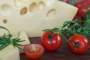 Big piece of cheese with holes lying with fresh twigs rosemary and cherry tomatoes on a wooden cutting board background.