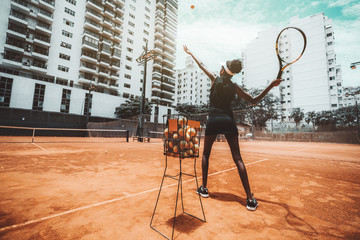 Wide-angle view from behind of a young slim biracial woman aiming a blow before hit a tennis ball during a sport training or warming-up on an outdoor tennis court, a metal basket with balls near her