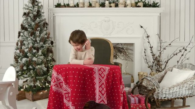 Cute little boy calling Santa while sitting on a big armchair at home over Chirstmas tree background