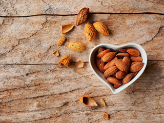 Raw almonds in heart shape bowl and broken nutshell top view on a rustic wooden table with copy space on the left side of the picture for healthy eating or vegan food concept.