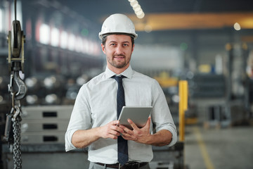 Portrait of confident handsome technical engineer in hardhat using tablet in industrial shop of...