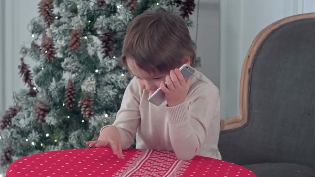 Cute little boy calling Santa while sitting on a big armchair at home over Chirstmas tree background