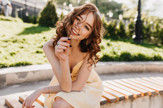 Cute female model with wavy red hair sitting on park bench. Outdoor photo of blissful ginger girl in yellow dress posing on nature background.