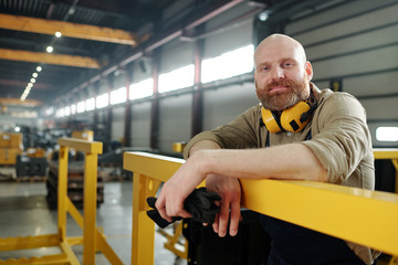 Portrait of content bald manual worker with beard holding work gloves and leaning on metal railings...